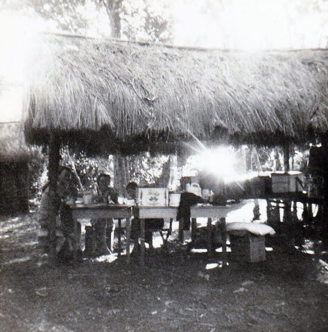 Open air thatch roof hut where Mary and Linda are sitting at a table, c 1972. ©KMC