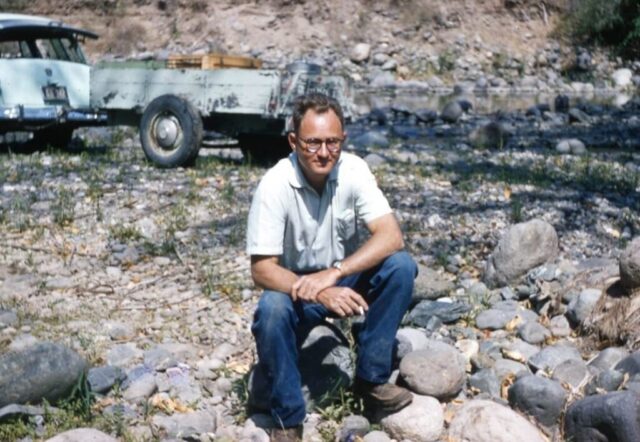 Kennedy M Crockett sits on a rock smoking a cigarette with the station wagon and trailer in the background. Río Los Plátanos, Guatemala, 1957. ©KMC