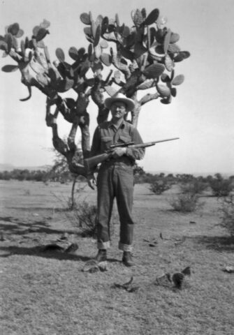 A man in hunting gear stands in front of a tall nopal cactus in a desert area in México, 1949.