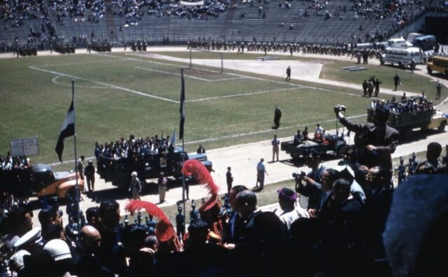 A parade of military vehicles and soldiers in a sports arena with flags and onlookers in the stadium seats. Inauguration of President General Miguel Ydígoras Fuentes, Guatemala City, Guatemala, 1958. ©KMC