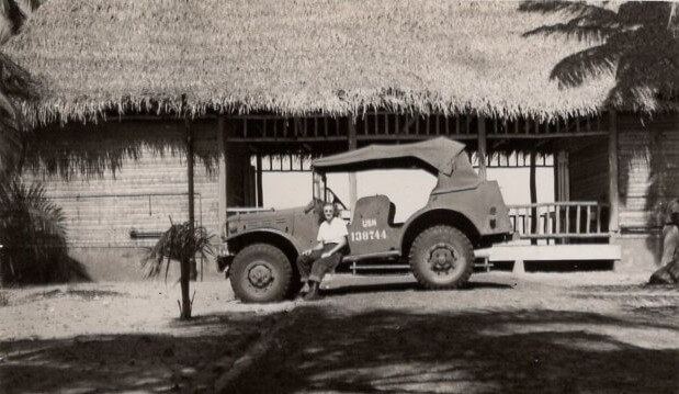 A young woman wearing sunglasses sits of the driver's side step of a jeep used during WWII with a thatch-roofed hut in the background. La Ceiba, Honduras, 1947. ©KMC