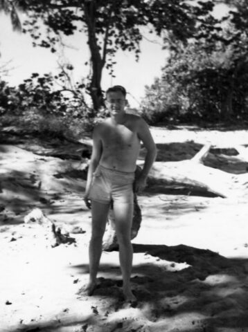 A barefoot young man in swimming trunks is leaning again a tree on the beach - Kennedy M Crockett, US Vice Consul, La Ceiba, Honduras, 1947. ©KMC