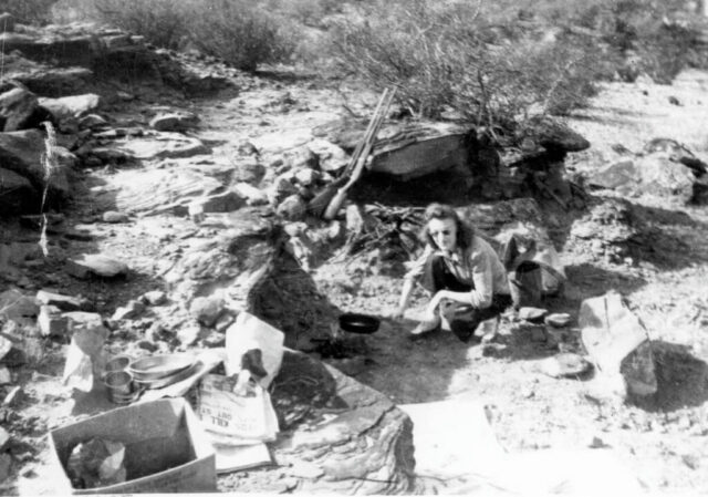 A young woman is placing a cast iron skillet on a campfire out in the country, c 1943-1944. ©KMC