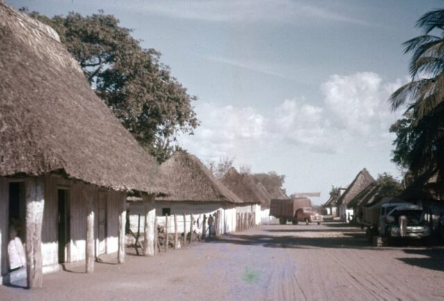 The dirt Pan-American Highway passes through the town of Sipacate, Guatemala, 1957. The wooden houses have white walls and thatched-roofs. ©KMC