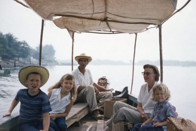 Three children and their mom ride down a wide river in a canvas-covered panga. The boatman steers the boat from the back. Iztapa, Guatemala, 1958. ©KMC