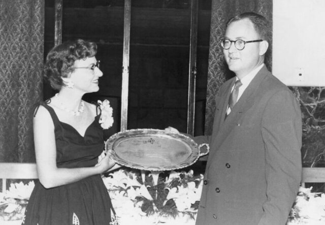 A woman and a man smile and stand holding a silver tray that was gifted to them at their going away party. Tampico, México, 1955. ©KMC