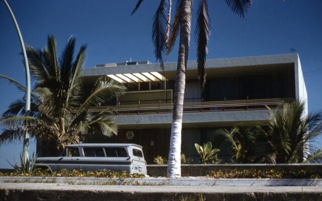 The Consulate's carryall is parked in front of a modern two-story building surrounded by palms and blue sky, the US Consulate in Mazatlán, México, 1961. ©KMC