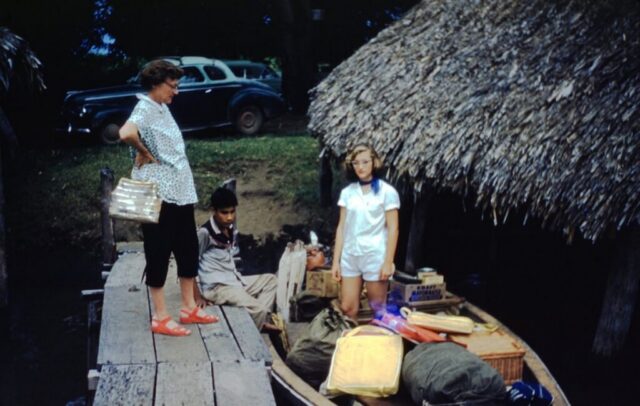 A woman waits on a wooden dock while her daughter stands in a small boat packed to the brim with camping gear and provisions. Embarcadero, Iztapa, Guatemala, 1958. ©KMC