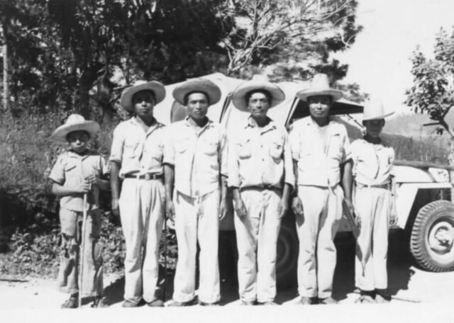 A line of indigenous men and boys dressed in white clothes and hats stand in front of a jeep in Honduras, 1946. ©KMC