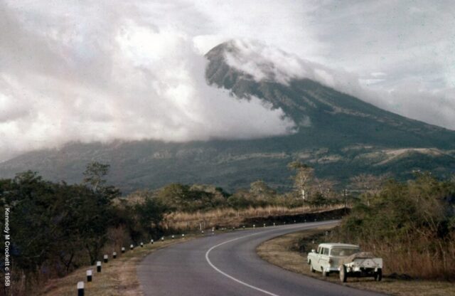A station wagon and trailer are parked alongside a paved road in front of a looming volcano. Volcán de Agua, Guatemala, 1960. ©KMC